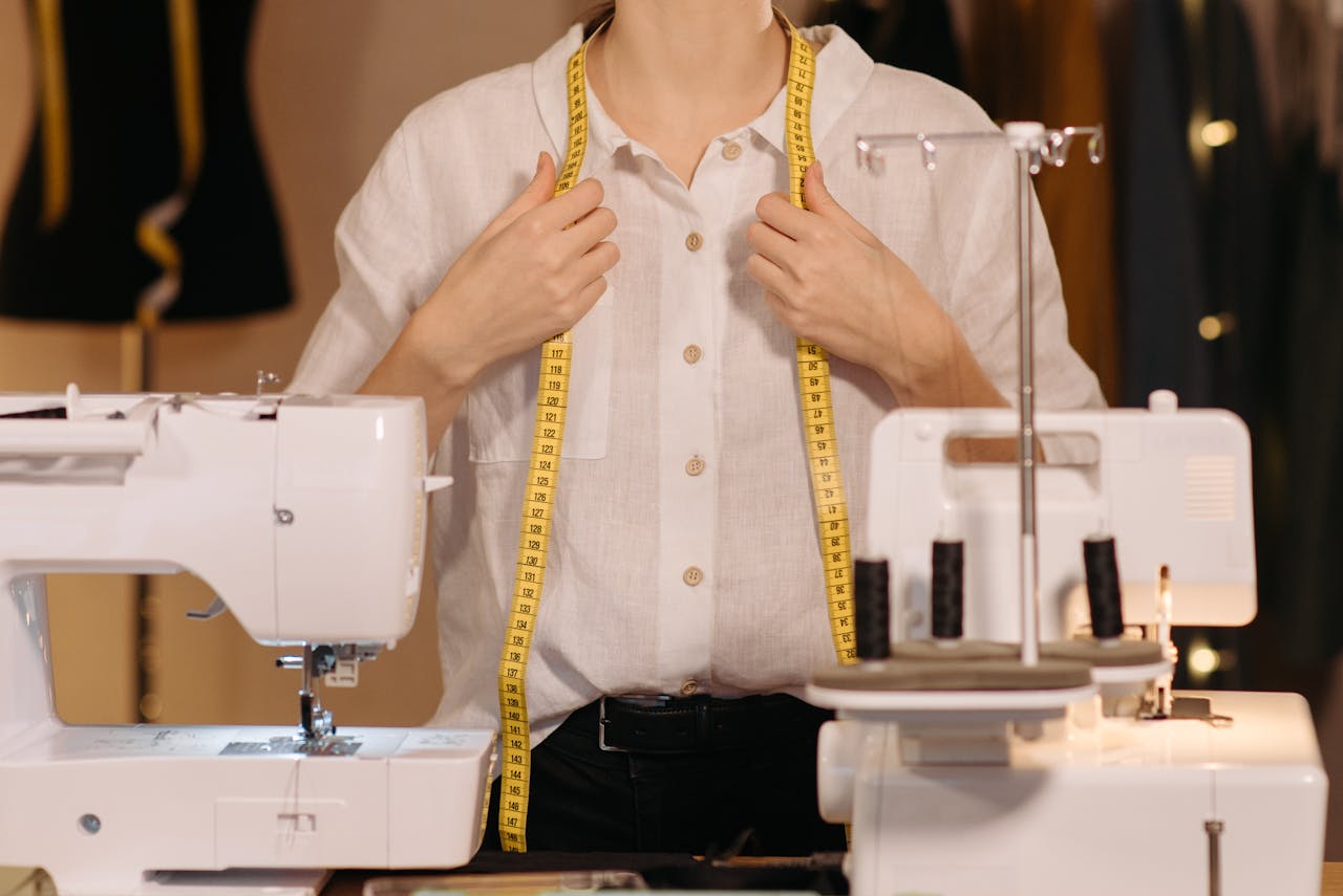 Tailor in a white shirt uses a measuring tape with sewing machines in a workspace.
